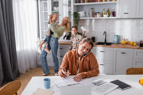 Man talking on mobile phone near blurred laptop and happy family playing on blurred background — Stock Photo