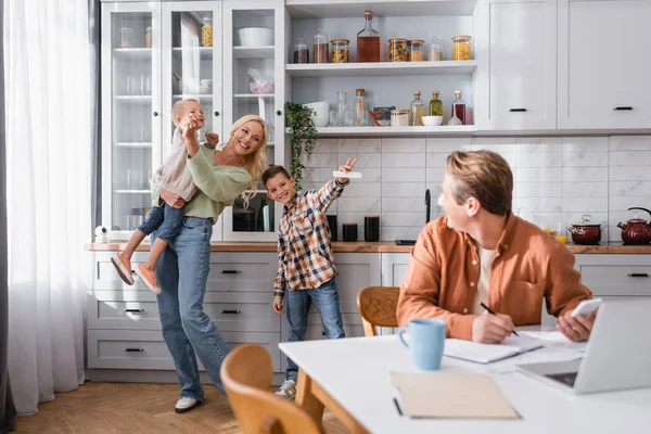 Cheerful woman having fun with sons near blurred husband working in kitchen — Stock Photo