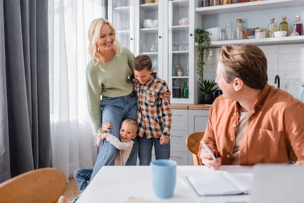 Fröhliche Frau umarmt Kinder neben Ehemann, der in Küche auf verschwommenem Vordergrund arbeitet — Stockfoto