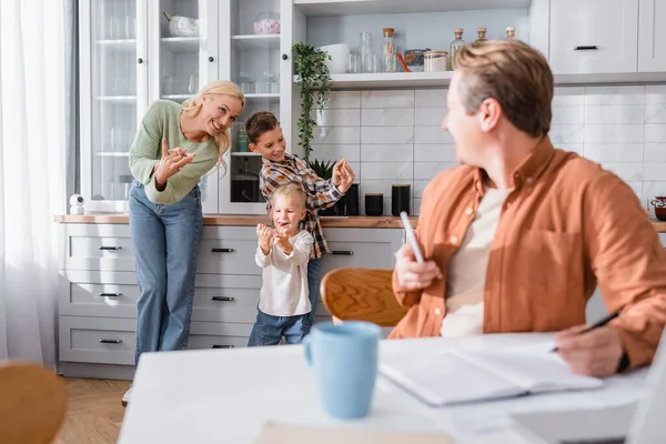 Cheerful woman playing in kitchen with sons near husband working on blurred foreground — Stock Photo