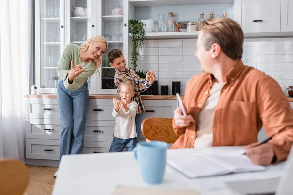 Fröhliche Frau spielt in der Küche mit Söhnen in der Nähe ihres Mannes und arbeitet an verschwommenem Vordergrund — Stockfoto