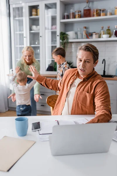 Busy man near laptop showing stop gesture to woman with kids playing in kitchen — Stock Photo