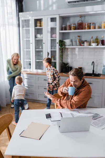 Homme avec tasse de thé pointant avec la main à la famille s'amuser dans la cuisine tout en travaillant près d'ordinateur portable flou — Photo de stock