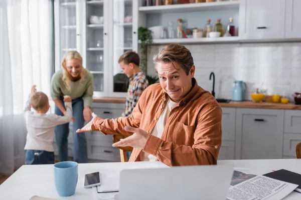 Découragé homme pointant vers la famille heureuse s'amuser dans la cuisine tout en travaillant près flou carnet — Photo de stock