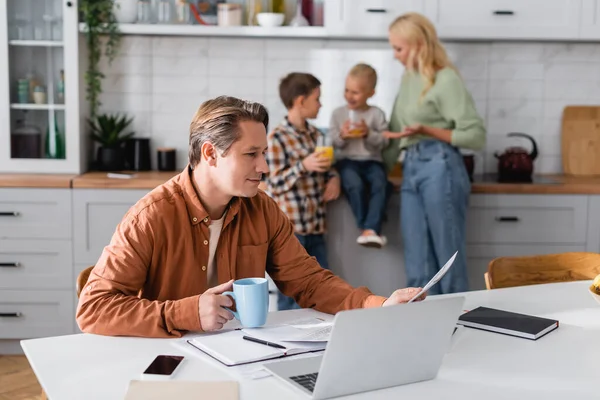 Mann mit Tasse Tee und Dokumenten arbeitet in Küche in der Nähe verschwommene Familie Orangensaft trinken — Stockfoto