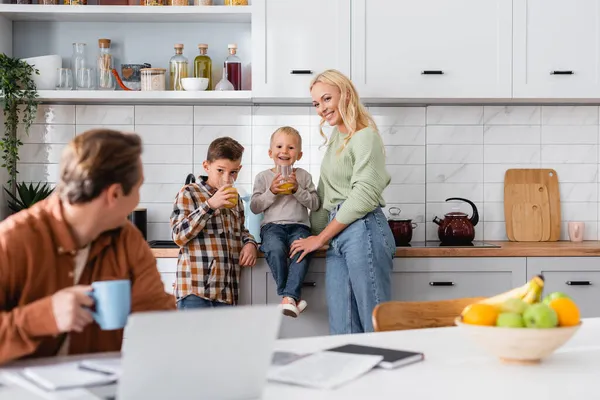 Kids drinking orange juice near happy mom and father freelancer working on blurred foreground — Stock Photo