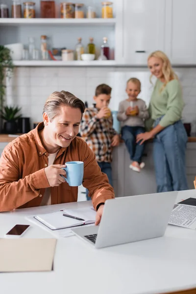 Lächelnder Mann mit Tasse Tee, der in der Küche in der Nähe von Laptop und Familie auf verschwommenem Hintergrund arbeitet — Stockfoto