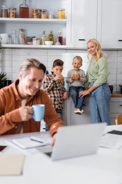 Glückliche Jungen trinken Orangensaft in der Nähe von Mama und Papa, die an verschwommenem Vordergrund arbeiten — Stockfoto