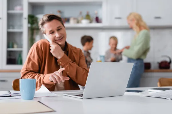 Homme parlant sur smartphone et pointant vers un ordinateur portable tout en travaillant près de la famille floue dans la cuisine — Photo de stock