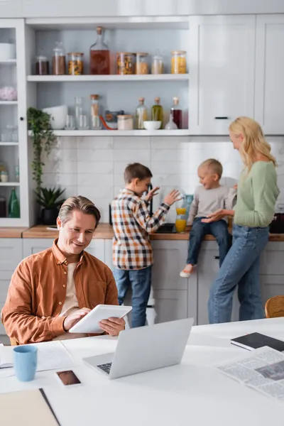 Homem sorridente com tablet digital trabalhando perto de laptop e família beber suco de laranja na cozinha em fundo borrado — Fotografia de Stock