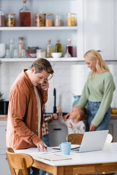 Freelancer falando no telefone celular enquanto trabalhava com documentos na cozinha perto de laptop e família turva — Fotografia de Stock