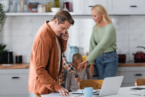 Positive man talking on mobile phone near documents, laptop and blurred family in kitchen — Stock Photo