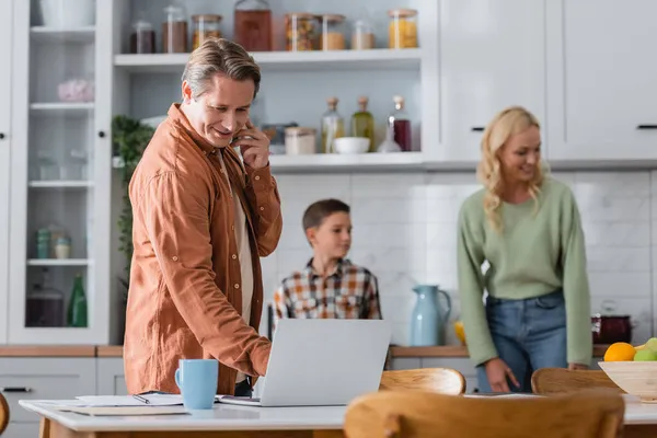 Blurred woman and son near man talking on smartphone while working near laptop in kichen — Stock Photo