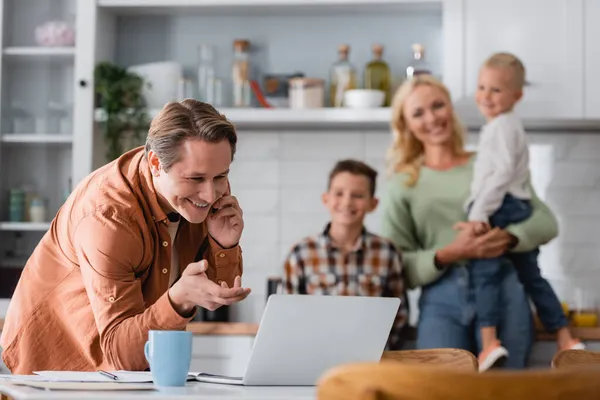 Hombre hablando en el teléfono inteligente mientras trabajaba cerca de la computadora portátil y la familia borrosa en la cocina - foto de stock