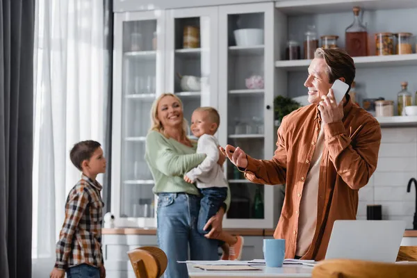 Homme souriant pointant avec la main tout en parlant sur smartphone près d'un ordinateur portable et la famille floue dans la cuisine — Photo de stock