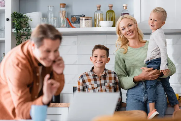 Vergrabener Mann zeigt Okay-Geste beim Gespräch auf Smartphone in der Nähe von Laptop und Familie in Küche — Stock Photo