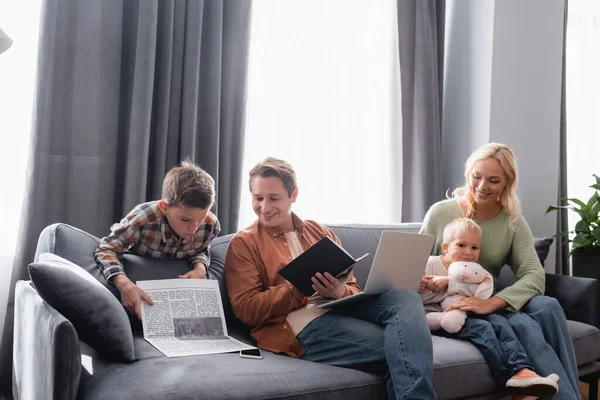 Boy reading newspaper near dad working with notebook and laptop near wife playing with kid on sofa — Stock Photo