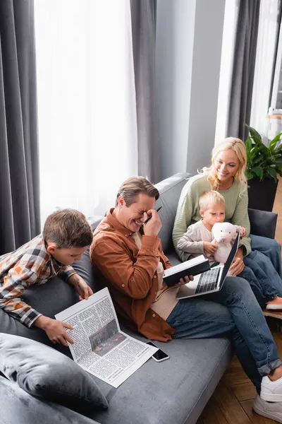 Menino lendo jornal perto de rir pai trabalhando com notebook e laptop perto da família sentado no sofá — Fotografia de Stock
