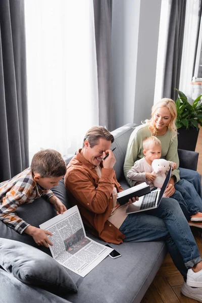 Boy reading newspaper near laughing father working with notebook and laptop near family sitting on sofa — Stock Photo