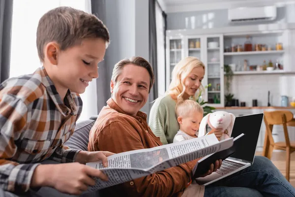 Boy reading newspaper near dad working with laptop and mom playing with brother on sofa — Stock Photo