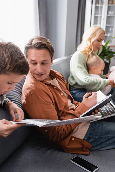 Boy pointing with finger while reading newspaper near father working on couch with with notebook and laptop — Stock Photo
