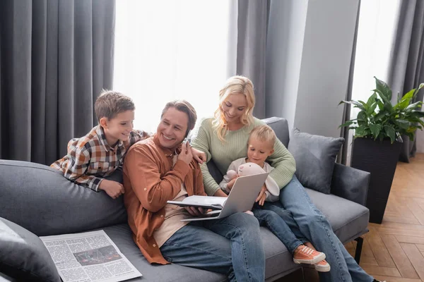 Улыбающийся мужчина разговаривает на смартфоне почему сидит на диване с ноутбуком рядом веселая семья — стоковое фото