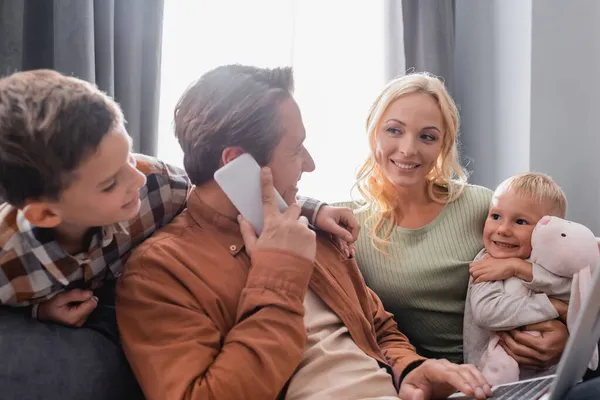 Hombre hablando en el teléfono móvil mientras trabaja en el ordenador portátil cerca de la familia feliz en el sofá en la sala de estar - foto de stock