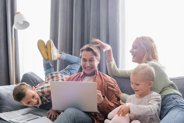 Alegre familia divirtiéndose en sofá cerca irritado hombre trabajando en portátil - foto de stock