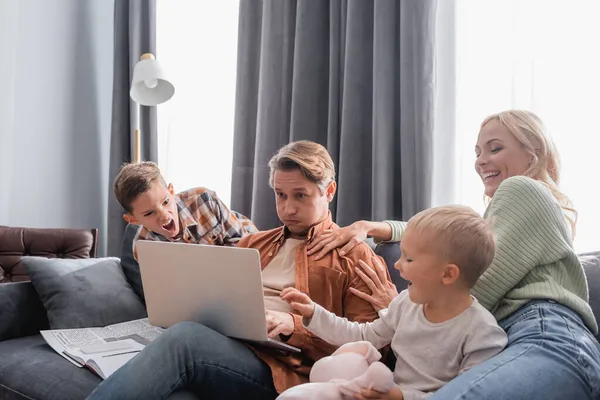 Geschäftiger und müder Mann bläst Wangen, während er am Laptop neben fröhlicher Frau und Söhnen arbeitet, die auf dem Sofa spielen — Stockfoto