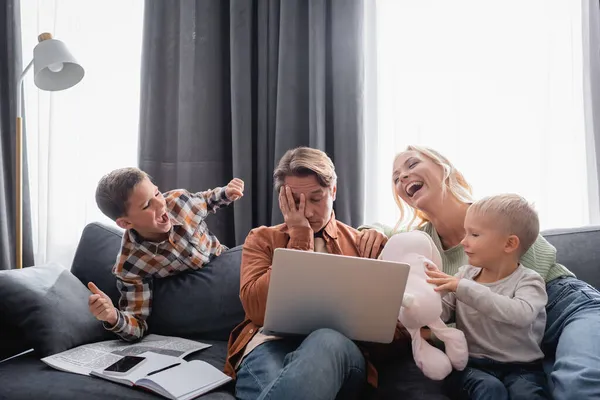 Tired man obscuring face with hand while working on laptop near family playing on sofa — Stock Photo