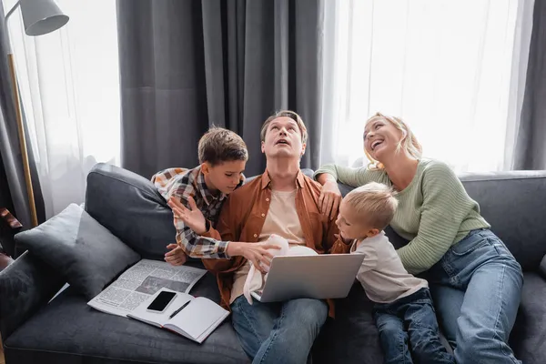 Busy and tired man with laptop trying to work near cheerful family playing fun on sofa — Stock Photo