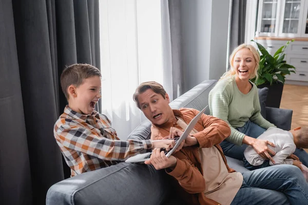 Ragazzo eccitato prendere laptop da occupato padre vicino a mamma ridere sul divano — Foto stock