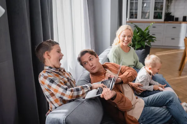 Ragazzo eccitato portare via computer portatile da papà seduto sul divano vicino moglie e bambino piccolo — Foto stock