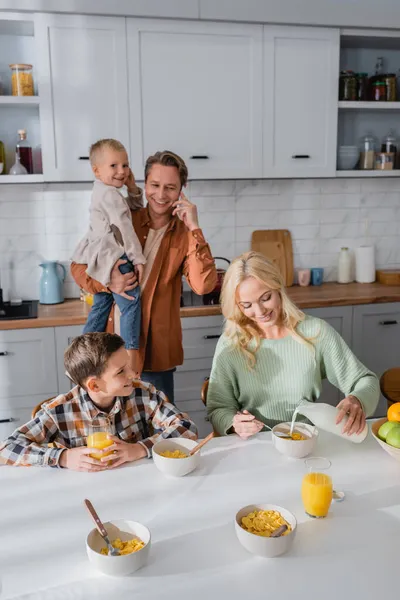 Женщина наливает молоко в хлопья кукурузы и муж разговаривает по смартфону во время завтрака с детьми — стоковое фото