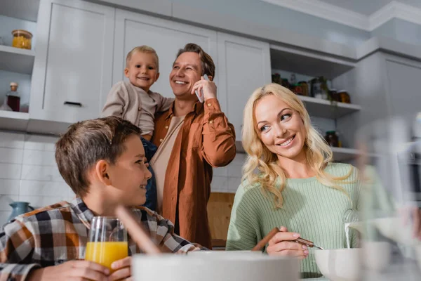 Smiling man holding kid and talking on smartphone near wife and son having breakfast in kitchen — Stock Photo