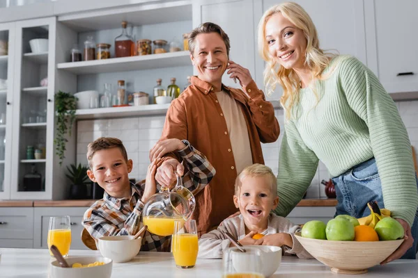 Smiling man talking on smartphone and pouring orange juice during breakfast with family — Stock Photo