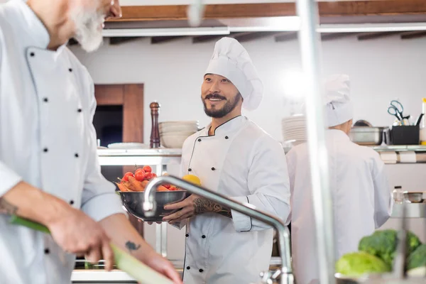 Sorrindo asiático chef segurando tigela com legumes perto colega na cozinha — Fotografia de Stock