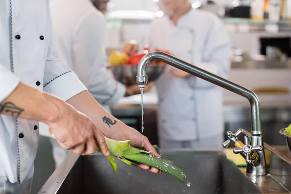 Обрізаний вид кухаря, що миє цибулю-порей біля розмитих колег на кухні — стокове фото