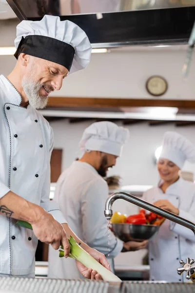 Sorrindo chef lavando alho-porro perto de colegas inter-raciais borrados na cozinha do restaurante — Fotografia de Stock