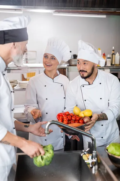 Chefs multiétnicos con verduras hablando con su colega cerca del fregadero en la cocina - foto de stock