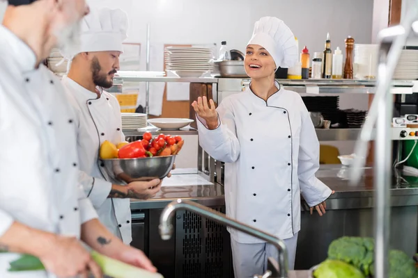 Chef sorrindo apontando com a mão perto de colegas inter-raciais com legumes na cozinha — Fotografia de Stock