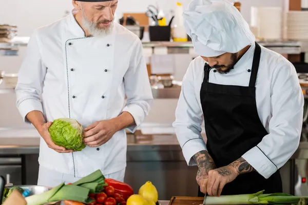 Chef asiático cortando puerro cerca de colega y verduras en cocina - foto de stock