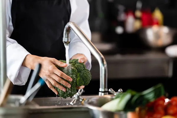 Ausgeschnittene Ansicht des Chefs, der Brokkoli in der Nähe von verschwommenem Gemüse in Küche wäscht — Stockfoto