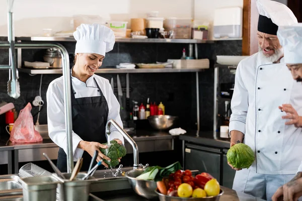 Smiling chef washing broccoli near interracial colleagues talking in restaurant kitchen — Stock Photo