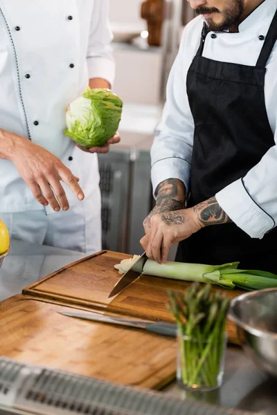 Cropped view of chef cutting leek near colleague with cabbage in kitchen — Stock Photo