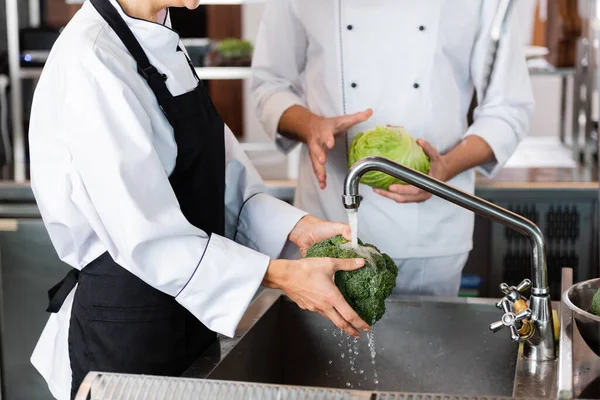 Cropped view of chef washing broccoli near colleague in kitchen — Stock Photo