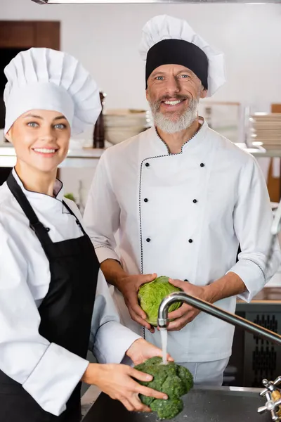 Chef sorrindo olhando para a câmera enquanto colega lavar brócolis na cozinha — Fotografia de Stock