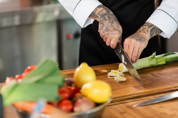 Cropped view of tattooed chef cutting leek near blurred vegetables in kitchen — Stock Photo