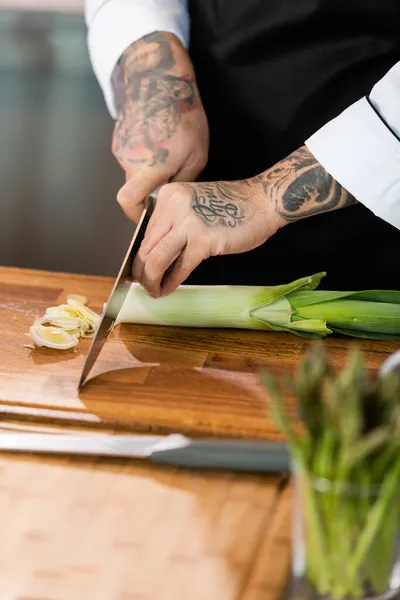 Vista cortada de chef tatuado cortando alho-porro perto de espargos em primeiro plano desfocado na cozinha — Fotografia de Stock