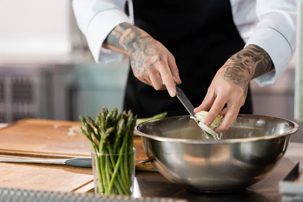 Обрезанный вид на татуированного шеф-повара, держащего нож и нарезанный лук-порей на кухне — стоковое фото